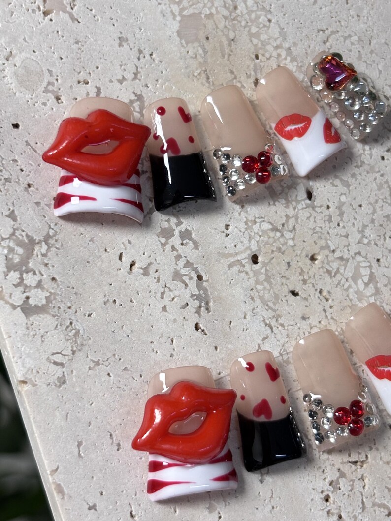 Pu&ograve; includere: Un set di unghie finte con vari design. Alcune unghie presentano labbra rosse, cuori rossi e una base nera. Altre sono decorate con strass e un top coat trasparente. Le unghie sono esposte su una superficie chiara e strutturata.