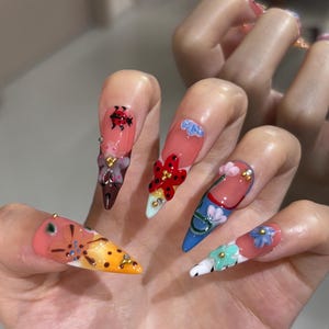 May include: Long, pointed artificial nails featuring a variety of colorful designs including a red ladybug, small blue and pink flowers, and abstract patterns in orange, yellow, blue, and white, accented with tiny gold beads and clear rhinestones.