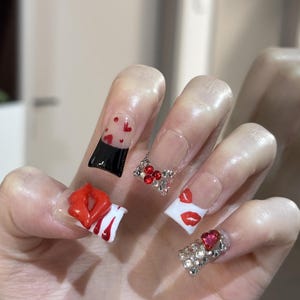 Pu&ograve; includere: Una mano con unghie acriliche lunghe e squadrate. Le unghie presentano vari disegni, tra cui labbra rosse, cuori e strass. Alcune unghie sono trasparenti con dettagli di cuori rossi, altre sono bianche con stampe di labbra rosse.