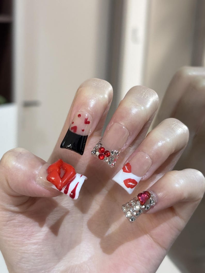 Pu&ograve; includere: Una mano con unghie acriliche lunghe e squadrate. Le unghie presentano vari disegni, tra cui labbra rosse, cuori e strass. Alcune unghie sono trasparenti con dettagli di cuori rossi, altre sono bianche con stampe di labbra rosse.
