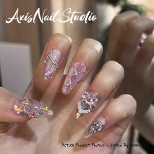 Puede incluir: Primer plano de una mano con uñas largas y decoradas. Las uñas presentan varios diseños, incluyendo purpurina, pedrería y patrones intrincados. El texto "Axis Nail Studio" está en la parte superior y "Actual Product Photos - Taken By Seller" en la parte inferior.
