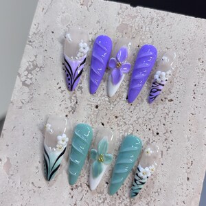 May include: A set of ten press-on nails featuring two distinct colour palettes of purple and teal. Each nail displays a mix of 3D floral accents, cable knit textures, and hand-painted black line art on a clear base with white tips.