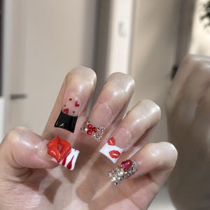 Pu&ograve; includere: Una mano che mostra unghie acriliche con vari design. Un'unghia presenta un design a labbra rosse, un'altra un design nero e rosso, e altre sono decorate con strass e dettagli a cuore. Le unghie sono di forma quadrata e hanno una base trasparente.