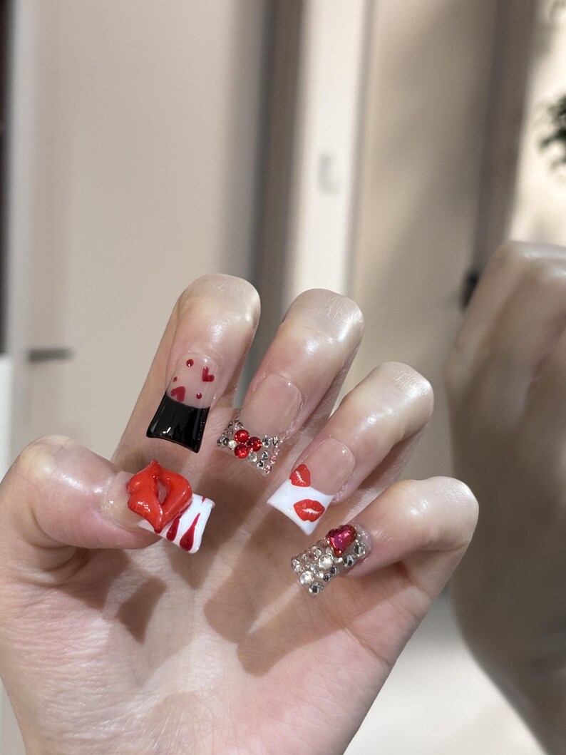 Pu&ograve; includere: Una mano che mostra unghie acriliche con vari design. Un'unghia presenta un design a labbra rosse, un'altra un design nero e rosso, e altre sono decorate con strass e dettagli a cuore. Le unghie sono di forma quadrata e hanno una base trasparente.