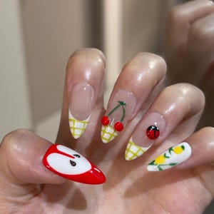 May include: Almond-shaped nails featuring a variety of fruit-themed nail art. Designs include a red apple slice, a yellow checkered pattern with cherries, a ladybug on a checkered tip, and yellow lemons with green leaves on a white base.
