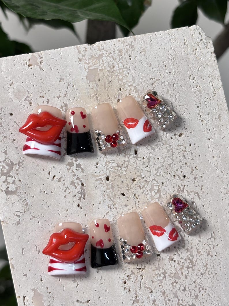 Pu&ograve; includere: Un set di unghie finte con vari disegni. Un'unghia presenta un design a labbra rosse 3D, mentre altre hanno accenti a cuore rosso, stampe di labbra rosse e strass. Le unghie sono di varie forme e dimensioni.