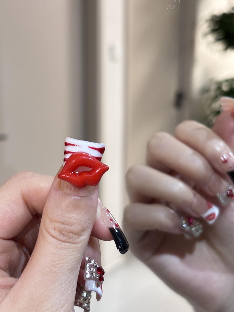 Pu&ograve; includere: Primo piano di una mano con unghie lunghe e decorate. Un'unghia presenta un design a labbra rosse, un'altra un gradiente nero e rosso, e altre sono decorate con strass e dettagli bianchi. Le unghie sono un'audace dichiarazione di moda.