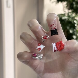 Pu&ograve; includere: Una mano che mostra unghie acriliche con vari disegni. Un'unghia presenta una punta nera con cuori rossi, mentre altre mostrano labbra rosse, strass e un design a labbra rosse 3D. Le unghie sono trasparenti.