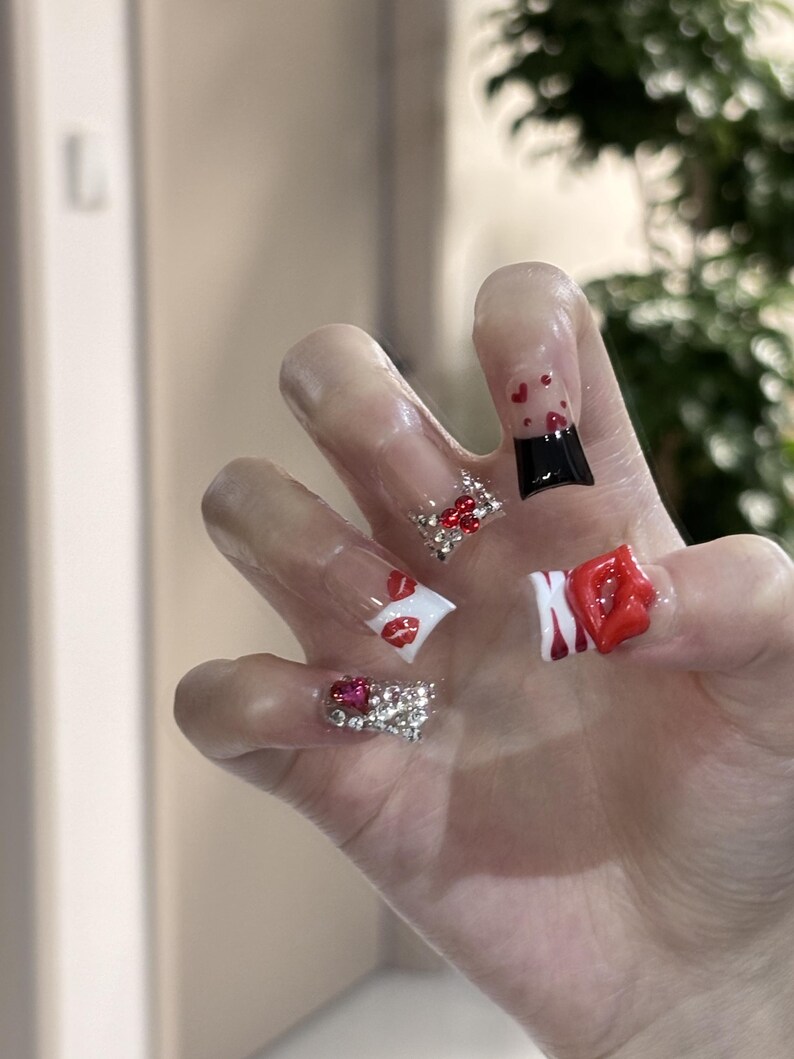 Pu&ograve; includere: Una mano che mostra unghie acriliche con vari disegni. Un'unghia presenta una punta nera con cuori rossi, mentre altre mostrano labbra rosse, strass e un design a labbra rosse 3D. Le unghie sono trasparenti.