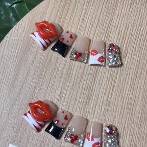 Pu&ograve; includere: Un set di unghie finte con vari disegni. Alcune unghie presentano labbra rosse, cuori e strass. Altre hanno uno schema di colori nero e nude. Le unghie sono disposte su una superficie di legno chiaro.