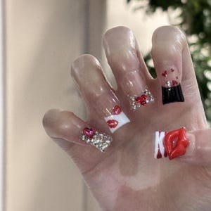 Pu&ograve; includere: Una mano con unghie lunghe e trasparenti in acrilico. Ogni unghia presenta disegni unici, tra cui labbra rosse, gemme a forma di cuore e un design nero e rosso. Alcune unghie sono decorate con strass scintillanti.