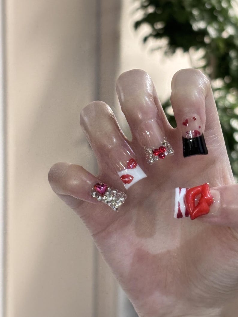Pu&ograve; includere: Una mano con unghie lunghe e trasparenti in acrilico. Ogni unghia presenta disegni unici, tra cui labbra rosse, gemme a forma di cuore e un design nero e rosso. Alcune unghie sono decorate con strass scintillanti.