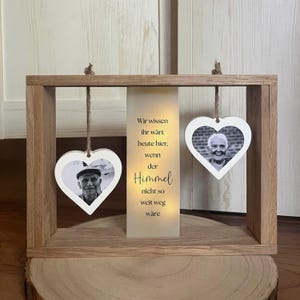 Könnte beinhalten: Ein rechteckiger Rahmen aus hellem Eichenholz mit zwei weißen, herzförmigen Fotorahmen, die an einer Schnur hängen. Dazwischen befindet sich ein beleuchtetes Paneel mit dem Text: Wir wissen ihr wärt heute hier, wenn der Himmel nicht so weit weg wäre.