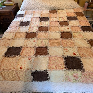 May include: A patchwork rag quilt featuring a grid of square fabric patches in shades of brown, cream, and soft pink. The quilt displays a variety of patterns including floral prints, polka dots, and textured solids with frayed, exposed seams.