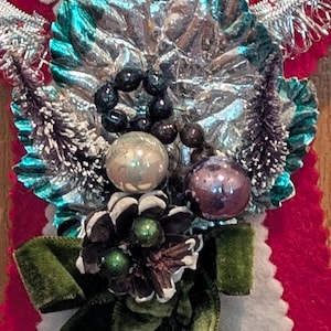 May include: A decorative ornament featuring a large, crinkled teal leaf, a cluster of black beads, a silver sphere, a purple sphere, a pinecone with green accents, and a dark green velvet ribbon, all mounted on a red and white felt backing.