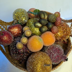 May include: A woven basket filled with assorted artificial fruits, including glitter-covered apples, oranges, and grapes in shades of gold, red, orange, and dark green. The decorative fruit pieces feature textured, sparkling surfaces and are arranged together in a round, light brown wicker container.