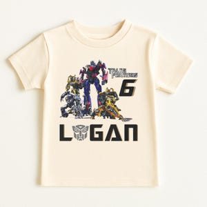 May include: Cream-colored short-sleeved t-shirt featuring a graphic of Transformers characters. The design includes the text "TRANSFORMERS 6" and the name "LOGAN" in black block letters. The Transformers logo is also present.