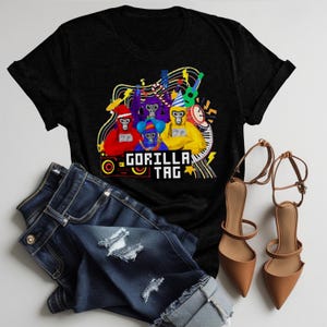 May include: Black t-shirt with a colorful graphic of cartoon monkeys playing musical instruments, with the text "GORILLA TAG". The shirt is paired with distressed blue jeans and brown heels.