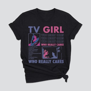 May include: Dark gray t-shirt with the text "TV GIRL" in purple and pink. The shirt features two graphic images and the text "WHO REALLY CARES" repeated multiple times. A list of song titles is also included.