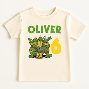 May include: A cream-colored t-shirt featuring a graphic of the Teenage Mutant Ninja Turtles. The name "OLIVER" is printed in green above the turtles, with a yellow number "6" to the right, indicating a birthday theme.