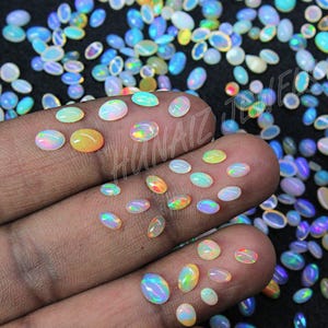 May include: A collection of oval-shaped opals in various sizes and colors, including white, yellow, blue, and green. Some opals are resting on a person's fingers, while others are scattered on a black surface. The opals display a play of color.