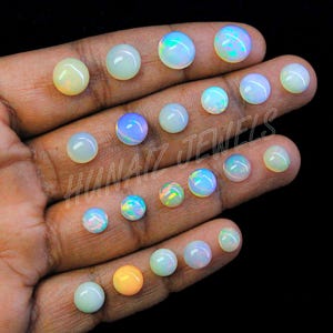 Puede incluir: Una mano muestra una colección de ópalos redondos de varios tamaños. Los ópalos exhiben una gama de colores, incluyendo blanco, amarillo, naranja y azules y verdes iridiscentes. Los ópalos están sobre un fondo oscuro.