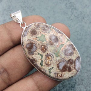 May include: Oval pendant with a patterned stone in shades of brown, beige, and green, set in a silver-coloured metal frame. The pendant has a loop at the top for a chain. The stone has a unique, organic pattern.