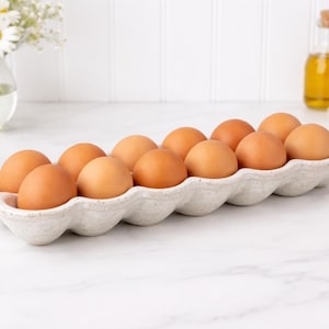 May include: A ceramic egg holder filled with a dozen brown eggs. The speckled, off-white holder sits on a white marble surface. A vase of white flowers and a bottle of oil are in the background.