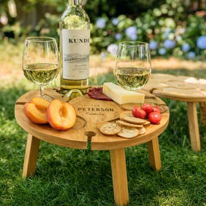 May include: A wooden picnic table with a bottle of wine, two wine glasses, and snacks. The table has slots for the wine bottle and glasses. The table is engraved with "The Peterson Family". Sliced peaches, crackers, cheese, and tomatoes are on the table.