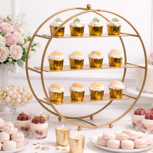 May include: A gold-colored, three-tiered cupcake stand holds cupcakes with white frosting and gold wrappers. The table is set with desserts, candles, and a floral arrangement, creating an elegant party setting.