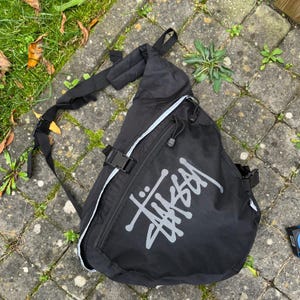 May include: Black sling bag with the white "Stussy" logo. The bag has an adjustable strap, a zippered main compartment, and a smaller front pocket. The bag is made of a durable material and features white trim.