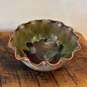 May include: A handmade ceramic bowl with a ruffled edge. The bowl features a blend of green, brown, and burgundy colors. The interior has a dark, glossy finish, and the rim is accented with a reddish-brown hue. The bowl is on a wooden surface.
