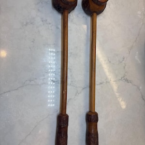 May include: Two wooden mallets with a dark brown finish. Each mallet has a round head with a small red accent and a long, slender handle. The handles are connected to a textured, carved base.