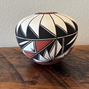 Puede incluir: Una olla de cerámica redonda, pintada a mano, con un diseño geométrico. La olla presenta un patrón de triángulos y formas negros, blancos y rojos. El borde es de color marrón oscuro. La olla está sobre una superficie de madera.