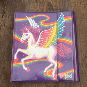 May include: A purple three-ring binder featuring a white winged horse with a pink mane and tail. The background is a dark purple starry sky with colorful rainbow streaks. The text Lisa Frank is printed in white with a rainbow outline at the bottom.