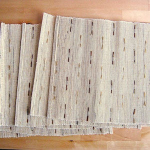 May include: A stack of beige rectangular placemats made of woven material. The placemats have a textured appearance with vertical brown and tan accents. The placemats are stacked on a light brown wooden surface.