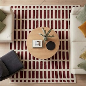 May include: Overhead view of a living room setup featuring a cream-colored rug with a repeating pattern of burgundy rectangles. A round wooden coffee table holds a plant, a book, and a black tray. Two white sofas and a dark gray armchair complete the scene.