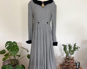May include: A black and white gingham midi dress with long sleeves, a black velvet collar and cuffs, and decorative buttons. The dress is displayed on a mannequin. A gold pendant necklace is also visible. The dress is a vintage style.