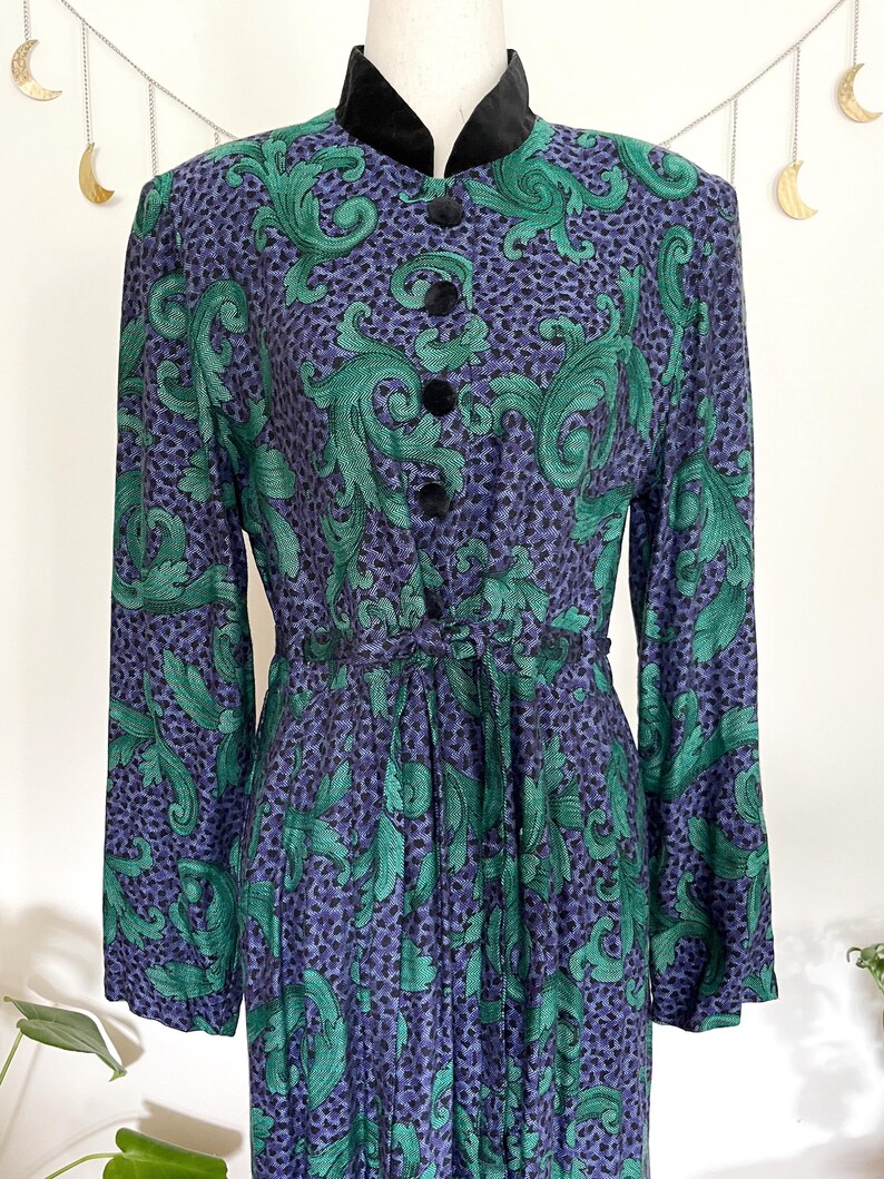 May include: A vintage dress with a high collar and long sleeves. The dress features a pattern of green leaves on a purple background with black dots. It has black velvet trim and buttons, and a matching tie belt.