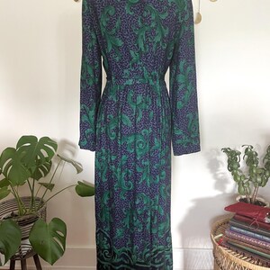 May include: A long-sleeved dress with a green and navy blue leaf pattern. The dress has a black collar and a fitted waist. The hemline features a decorative border. The dress is displayed on a mannequin.