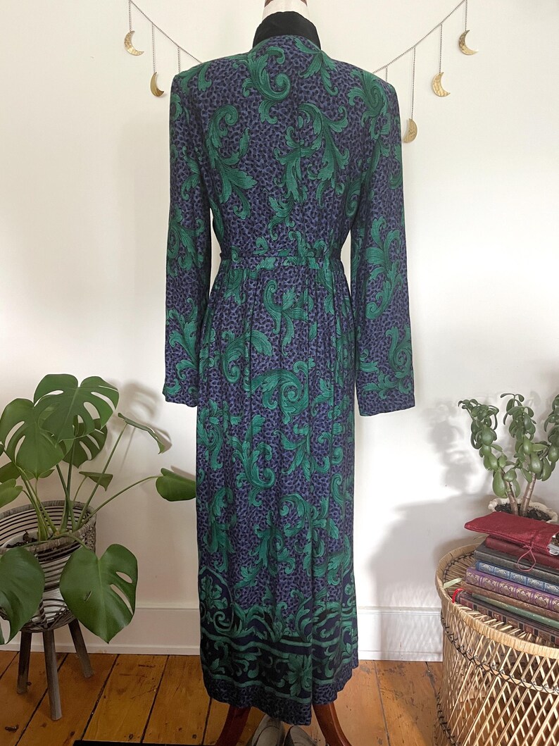 May include: A long-sleeved dress with a green and navy blue leaf pattern. The dress has a black collar and a fitted waist. The hemline features a decorative border. The dress is displayed on a mannequin.