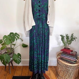 May include: A long, patterned dress in green and purple, worn with a cream cardigan and black boots. The dress features a paisley design and is accessorized with a gold pendant necklace. The outfit is displayed on a mannequin.