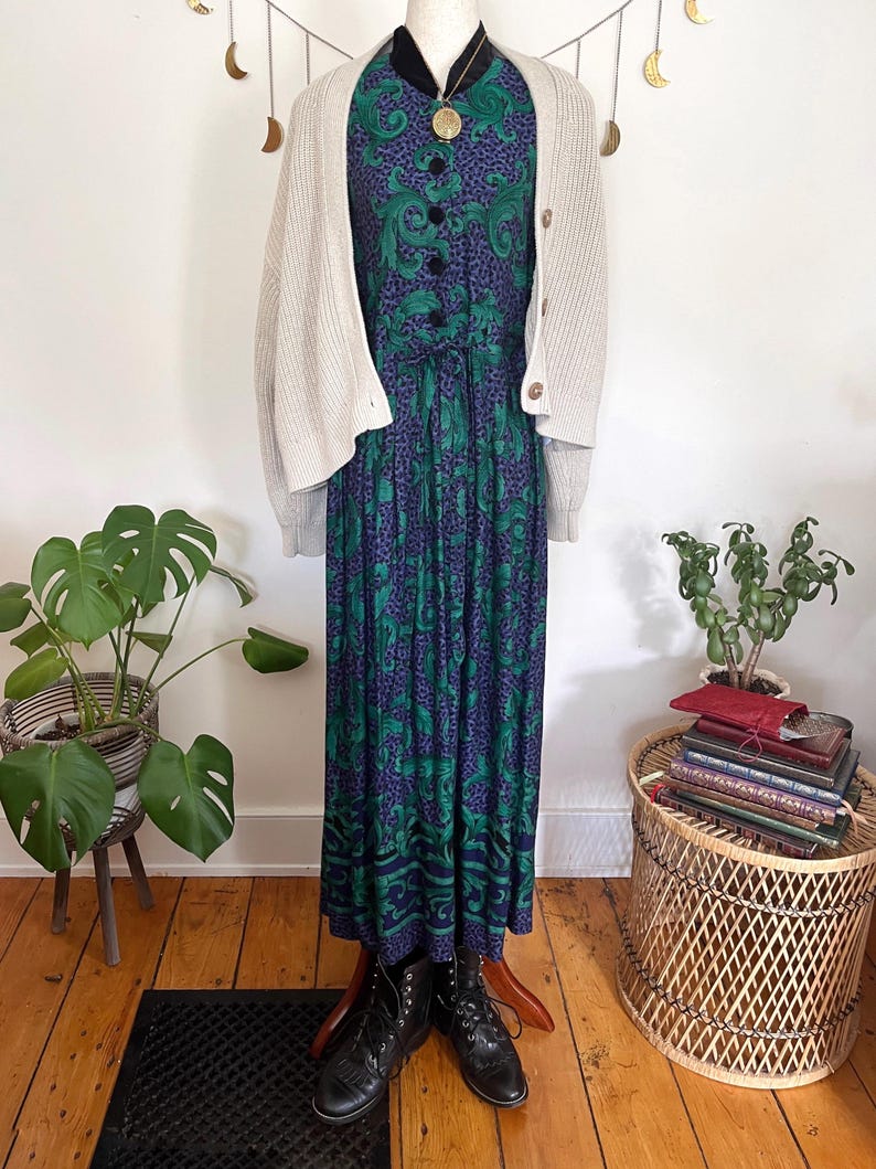 May include: A long, patterned dress in green and purple, worn with a cream cardigan and black boots. The dress features a paisley design and is accessorized with a gold pendant necklace. The outfit is displayed on a mannequin.