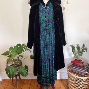 May include: A black velvet hooded coat over a patterned dress with green and purple accents. The dress is buttoned down the front and paired with grey buckle shoes. The outfit is displayed on a mannequin.