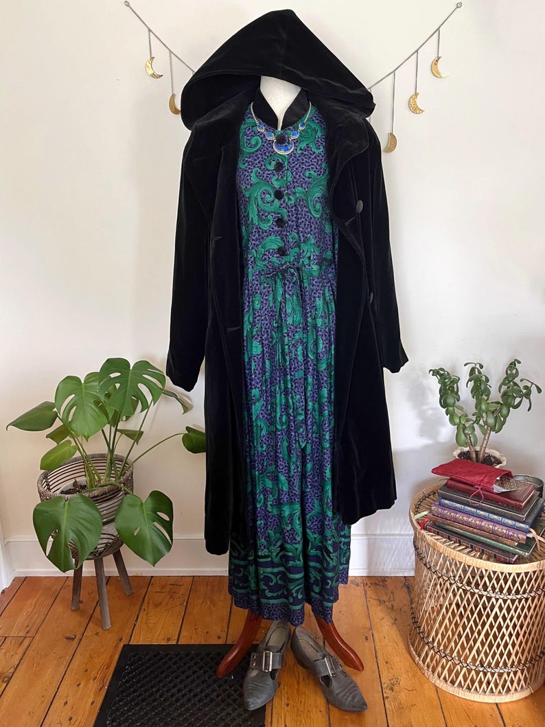 May include: A black velvet hooded coat over a patterned dress with green and purple accents. The dress is buttoned down the front and paired with grey buckle shoes. The outfit is displayed on a mannequin.