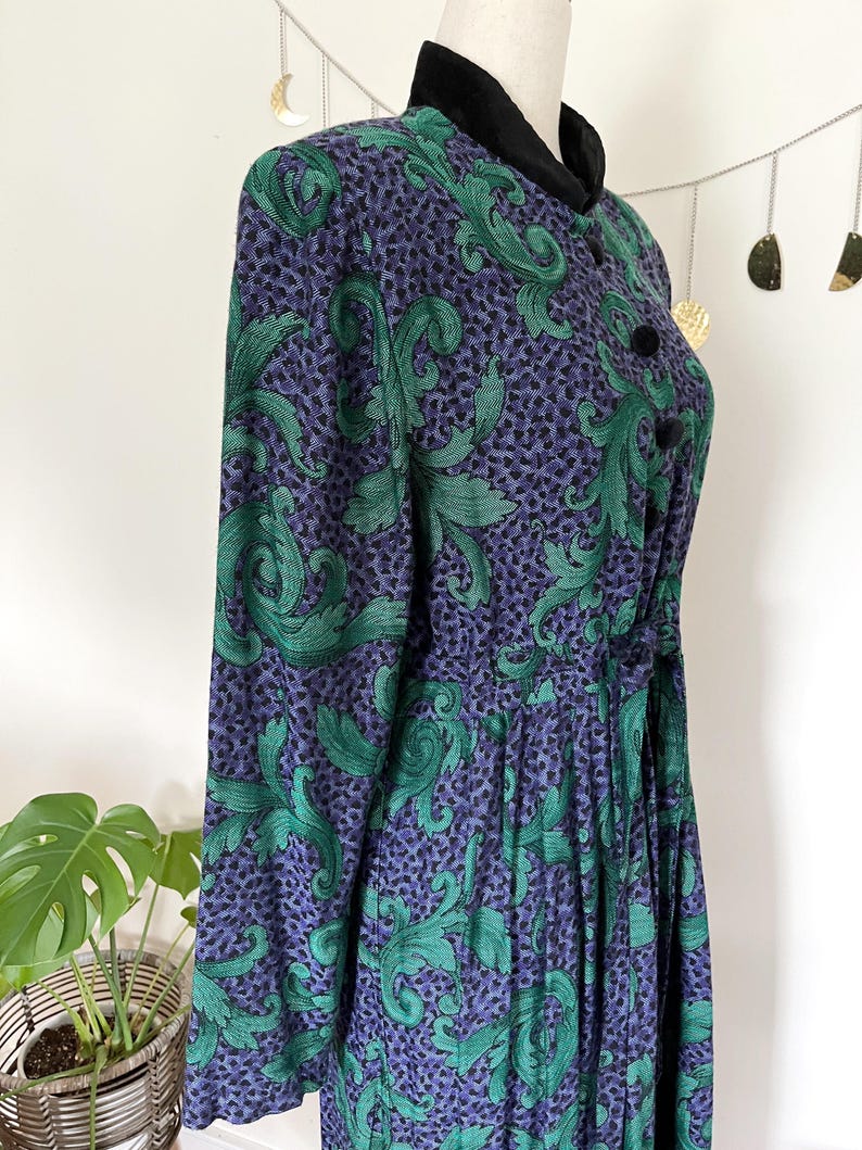 May include: A vintage dress with a high collar and long sleeves. The dress features a pattern of green leaves on a purple background. It has black buttons and a black velvet collar. The dress is displayed on a mannequin.