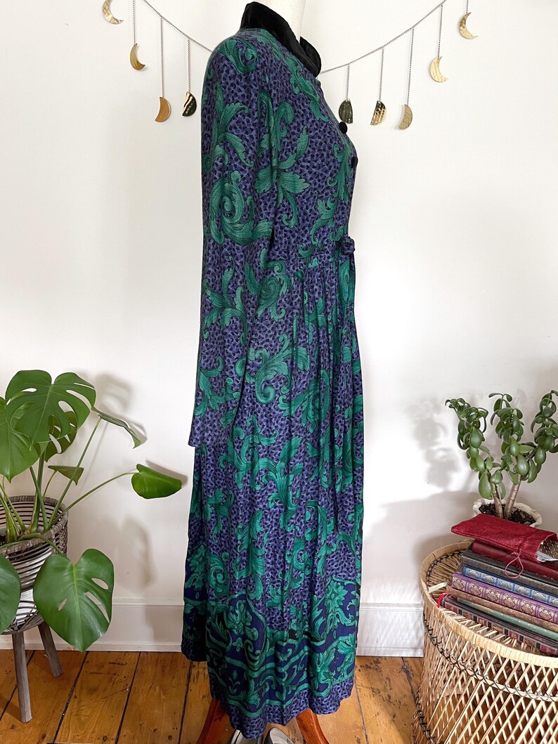 May include: A long, vintage dress with a dark blue and green floral pattern. The dress has a black velvet collar and buttons down the front. The skirt is pleated and reaches the ankles. The dress is displayed on a mannequin.