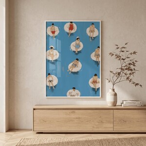 May include: A framed art print featuring a vibrant blue pool scene with people floating on white inner tubes. The people are wearing various swimsuits. The artwork is displayed on a light-colored wall above a wooden cabinet.