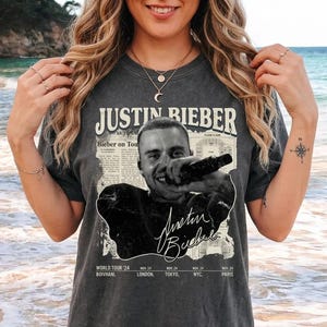 Puede incluir: Camiseta gris oscuro con una imagen en blanco y negro de Justin Bieber. La camiseta incluye el texto "JUSTIN BIEBER" y fechas de gira de 2024 en varias ciudades.