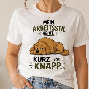 Könnte beinhalten: Weißes T-Shirt mit einer Cartoon-Hundeillustration und deutschem Text. Der Hund liegt, mit dem Text "MEIN ARBEITSSTIL HEIBT: KURZ = VOR = KNAPP." in Grün gedruckt. Das T-Shirt wird von einer Person in blauen Jeans getragen.